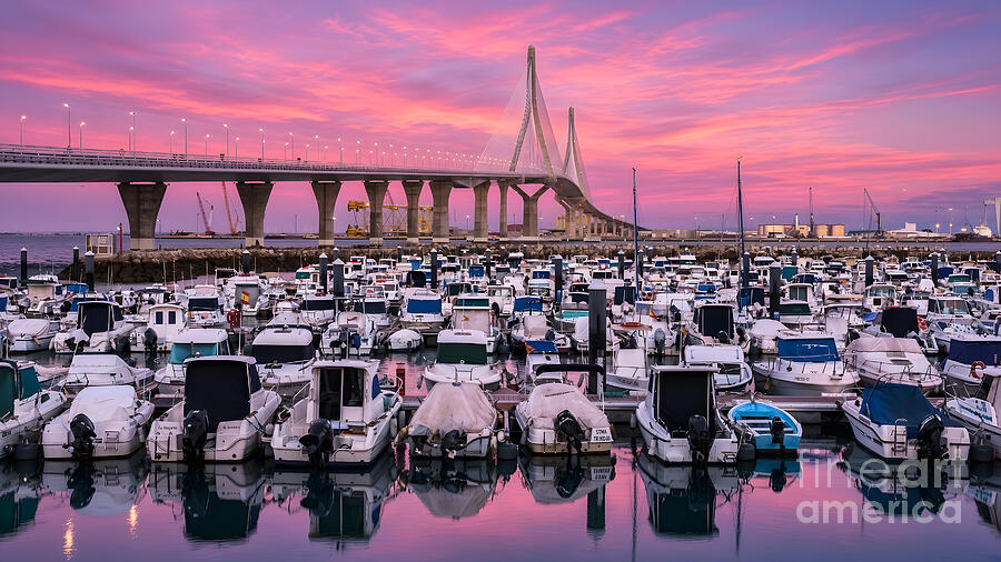 Rows of small boats moored in a marina on the Bay of Cadiz, with the 1812 Constitution Bridge silhouetted against a pink and violet sunset