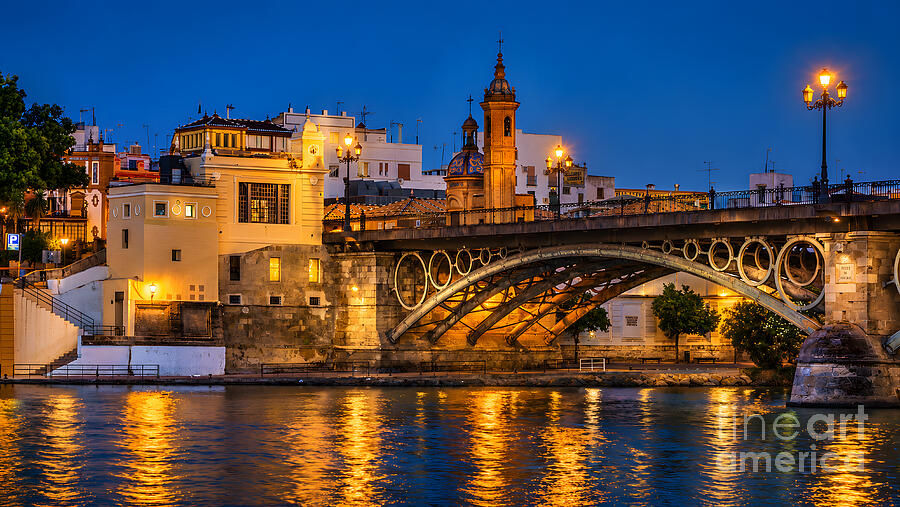 The Triana Bridge (Puente de Isabel II) in Seville at dusk, with lamps lit on the iron arch, the tower and dome of the Capilla del Carmen, and golden reflections in the river