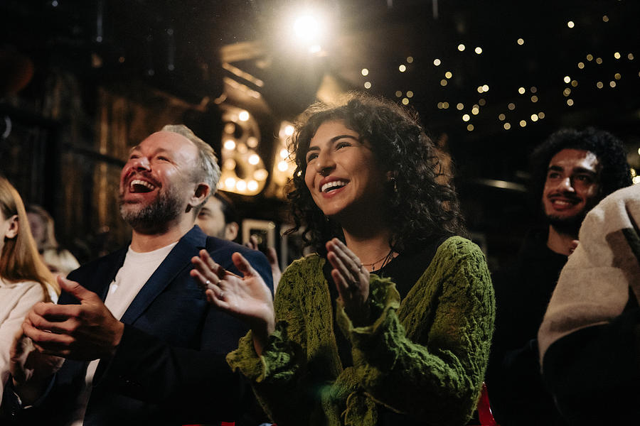 Happy audience laughing and clapping while watching comedy stage show in illuminated theater Photograph by Maskot