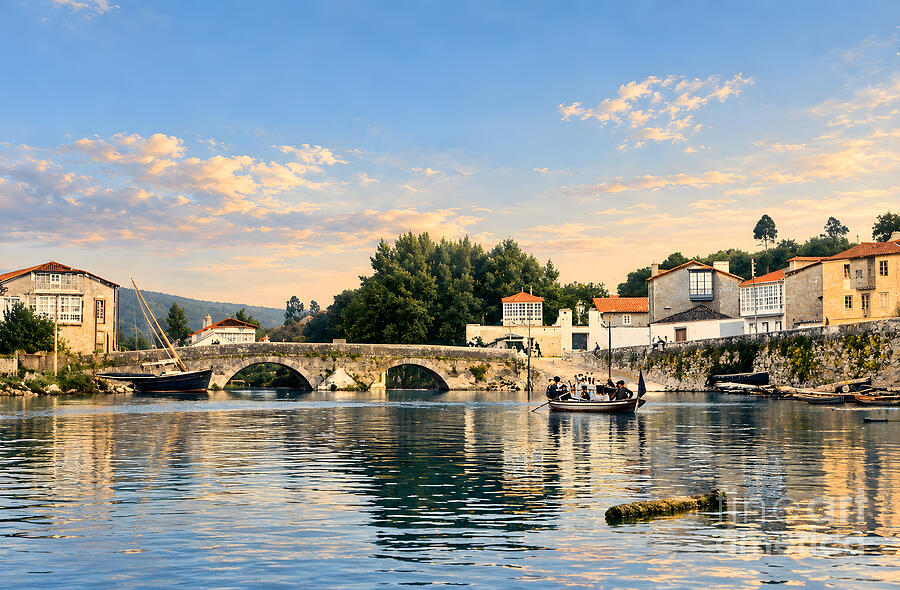 The Xubia (Jubia) bridge between Neda and Naron, Galicia, in 1906: the original postcard photograph, restored, with a boat of men on the calm estuary, the stone bridge and riverside houses in warm evening light