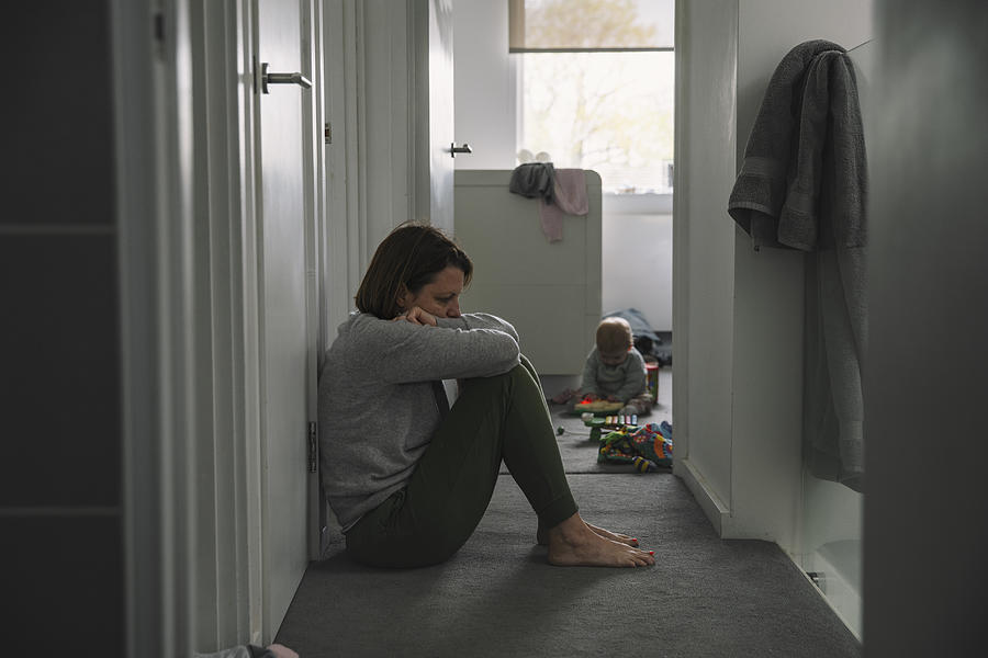Mature mother sitting on landing floor whilst her baby plays in the background - post natal depression Photograph by Justin Paget