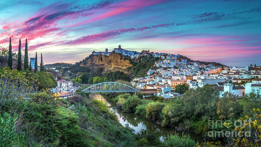 Our own photograph of Arcos de la Frontera at dusk: the white town on its cliff, the Guadalete river and the iron bridge under a pink and violet sky